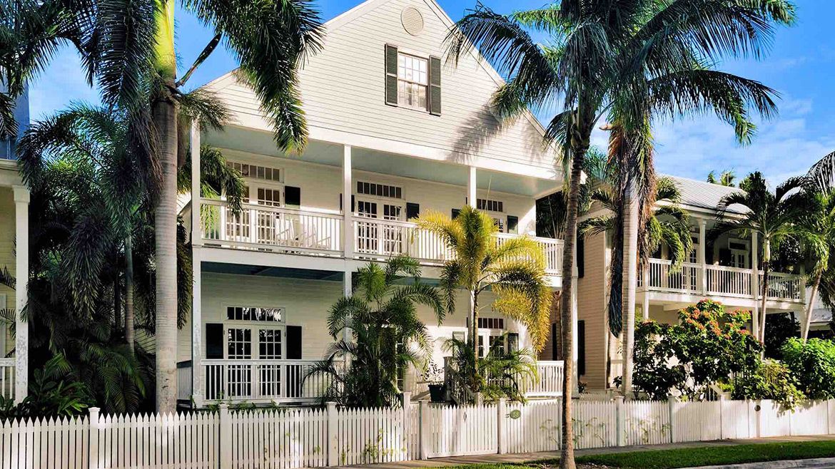A classic two-story white colonial house with double-stacked porches framed by tall palm trees. The home features traditional shuttered windows and is enclosed by a white picket fence, conveying a sense of elegant, tropical living.
