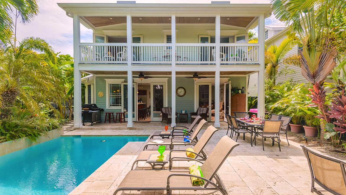 A tropical backyard oasis with a rectangular pool surrounded by sun loungers and lush greenery. The two-story house features a light blue siding with white trim, a spacious porch, outdoor ceiling fans, and an open dining area visible in the background