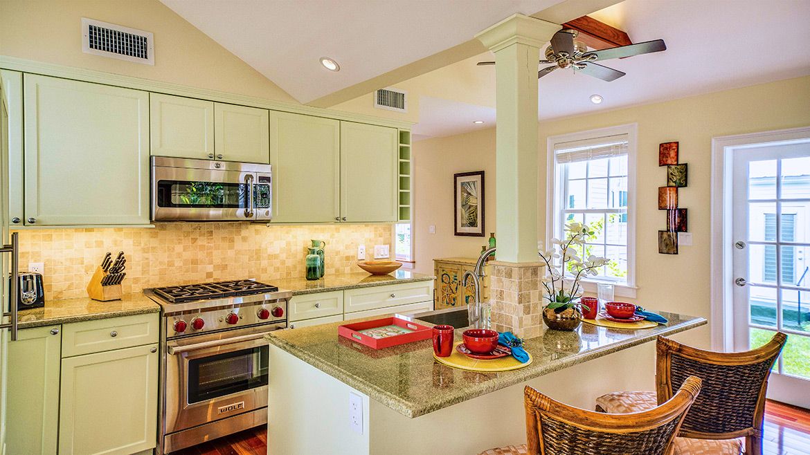 A well-lit, modern kitchen interior featuring pale green cabinetry, granite countertops, stainless steel appliances including a gas stove and microwave, and a kitchen island with red and blue bowls on top. The room has a beige tile backsplash, hardwood flooring, and a small dining area with wicker chairs.