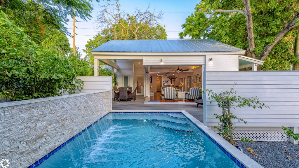 Photo of an outdoor pool at a residence in Key West, Florida. The rectangular pool is in the foreground which leads into the background that shows the open floor plan of a porch and living room area of the main house. 