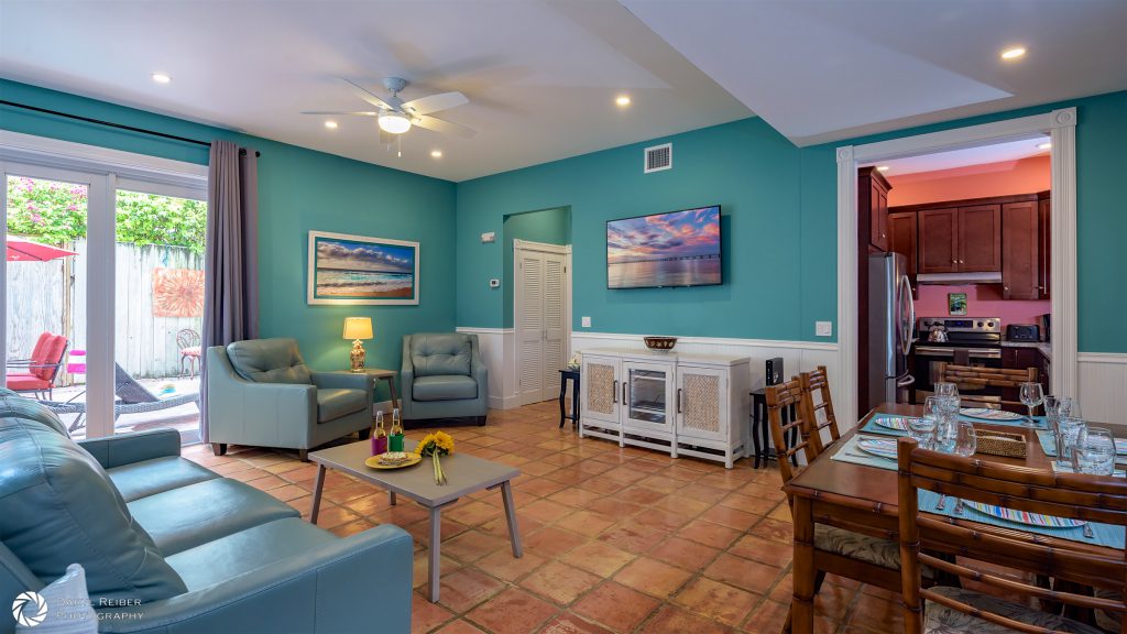 Photo of Key West real estate property named Good Vibrations. The living space features color matching couch and two chairs, a television mounted to the wall, and four chairs at a dining room table. 